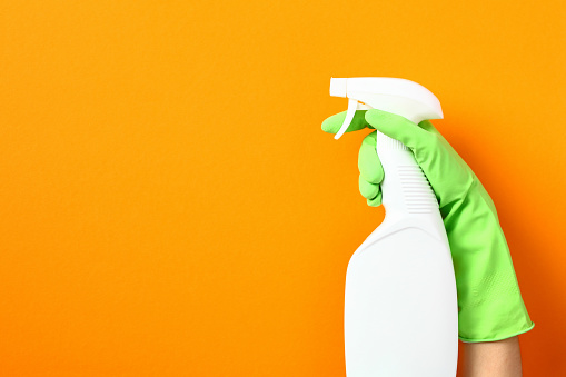 Disinfectant spray bottle mockup in female hand in green rubber glove over orange background. Kitchen cleaner product design.