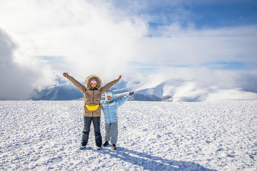 Two happy cheerful friendly girls in warm bright winter clothes are funny posing spreading their arms to the sides on a big snowy high mountain, against the backdrop of a cold sun covered with clouds