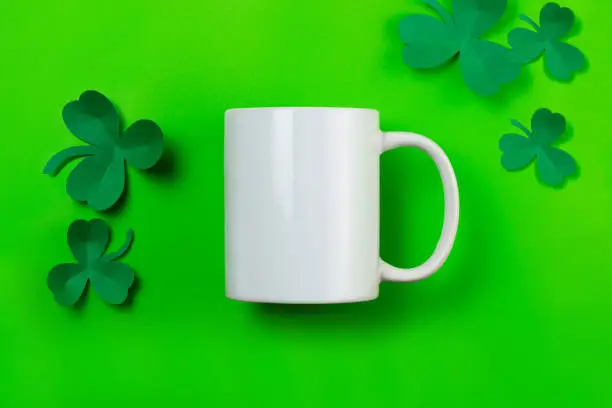 St. Patrick's Day empty white mug mockup. White coffee mug and paper clover shamrocks on green background