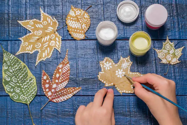 Girl paints patterns yellow maple leaf. Gouache, brush and various autumn leaves on a blue wooden table. Children's art project. Colorful Hand-painted on dry autumn leaves