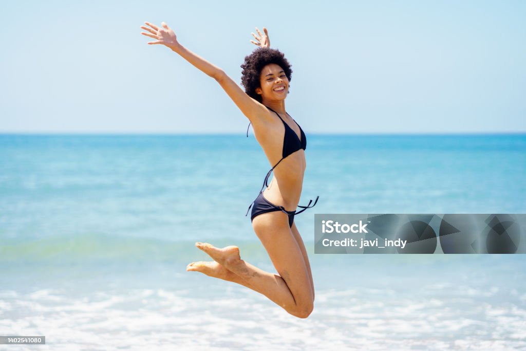 Happy black woman jumping near sea in summer Full body side view of cheerful African American female, traveler in bikini looking at camera, while jumping with raised arms against rippling sea on sunny day One Woman Only Stock Photo