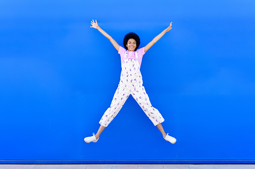Cheerful African American female looking at camera, while jumping with raised arms against a blue urban wall. Full body photograph
