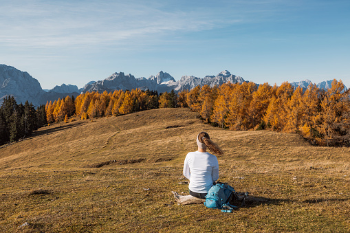 View from the back, young caucasian woman in autumn landscape, sitting on a grass field somewhere in the mountains, enjoying the view of autumn forest and mountain peaks in the background.