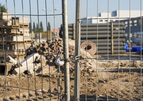 Close-up of metal fence doors which are closed with chain and padlock. In the background, out of focus, construction materals like pipes, bricks, sand, concrete. Far in the background a building and trees.