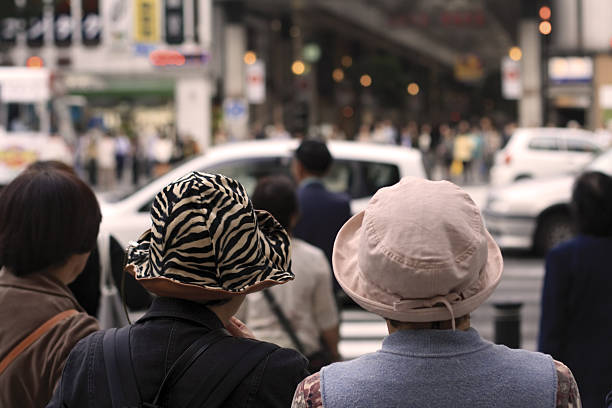 Very tipycal Japanese streetlife aspect with two senior women wearing hats waiting to cross the street in a crowdy dowtown. Selective focus on the hats.         