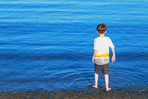 A boy steps carefully into the ocean at a rocky beach.