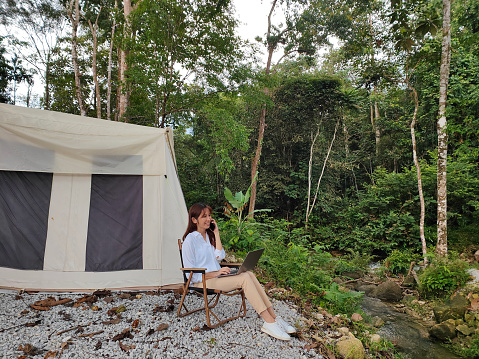 Young Asian woman working with smartphone and laptop during her camping vacation in the forest.