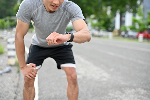 Exhausted asian male athlete hands on knee, checking his heart rate on smartwatch, taking a break after had a long running. cropped image