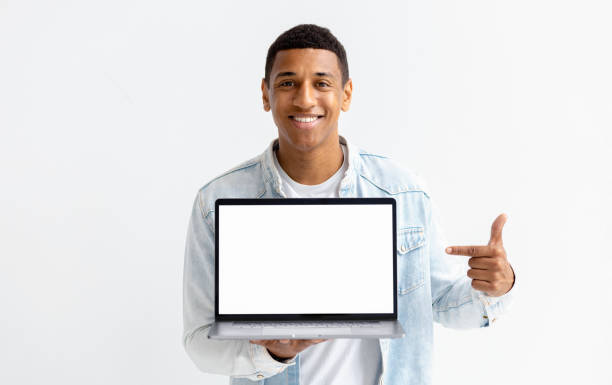 retrato de un joven afroamericano con una computadora portátil sobre fondo blanco. el hombre señala con el dedo la pantalla de una computadora portátil en blanco, mirando a la cámara y sonriendo - black sign holding vertical fotografías e imágenes de stock