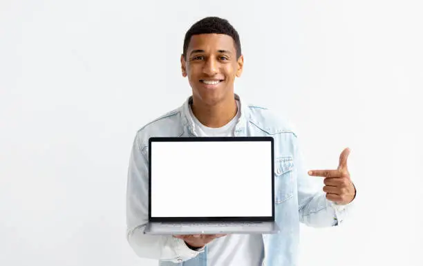 Photo of Portrait of young African American man with laptop on white background. Male points her finger at a blank laptop screen, looking at the camera and smiling