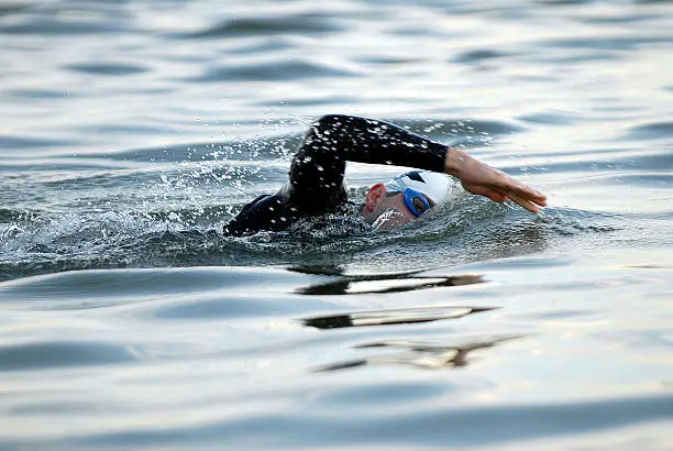 Male triathlete, mid-stroke, eye looking at camera through blue goggles in open water.  White swim cap.