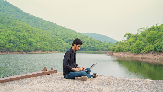 Young man working on a laptop and sitting in the nature, Workation and Digital Nomad concept.