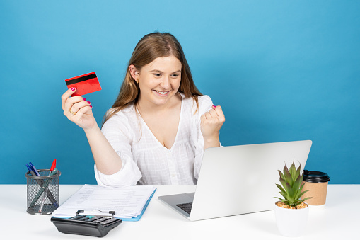 Young pretty office worker celebrating purchase and holding credit card isolated on blue background. Online shopping concept