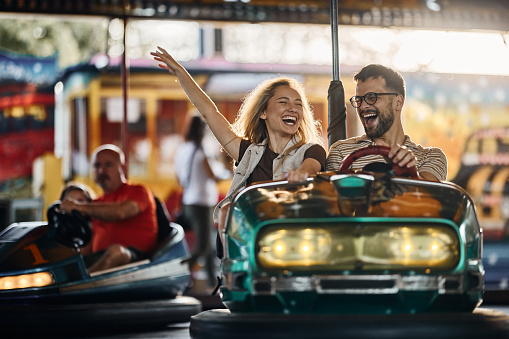 Young cheerful couple having fun in bumper car ride at amusement park.