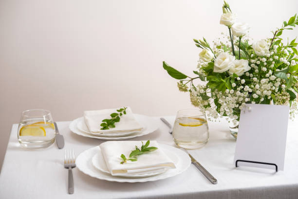 Mockup white blank space card for table number or menu. Wedding teble decoration with white flowers, glasses and white napkins. Elegantly decorated table at a wedding reception. Festive table setting. The wedding decor.