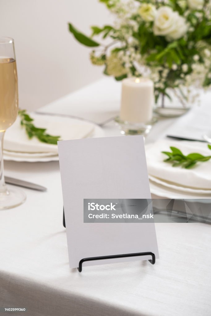 Mockup white blank space card for table number or menu. Wedding teble decoration with white flowers, glasses and white napkins. Elegantly decorated table at a wedding reception. Festive table setting. The wedding decor. Mockup white blank space card for table number or menu. Wedding table decoration with white flowers, glasses and white napkins. Elegantly decorated table at a wedding reception. Festive table setting. The wedding decor. Number Stock Photo