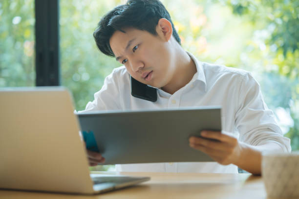 Asian man working busy while making phone call and using laptop at home stock photo