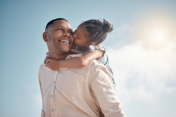 Smiling mixed race single father carrying little daughter on piggyback with copyspace. Adorable, happy, hispanic girl bonding with parent and kissing cheek on beach. Man and child enjoying free time stock photo