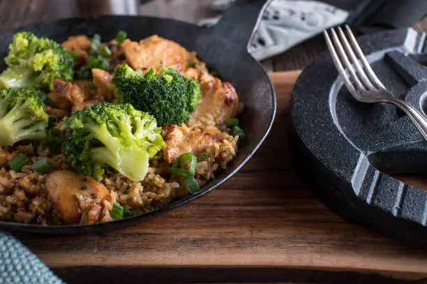 Healthy meal for muscle gain with seared chicken breast, whole grain rice and broccoli. Served in a rustic iron pan with weight plate on wooden table background. Closeup