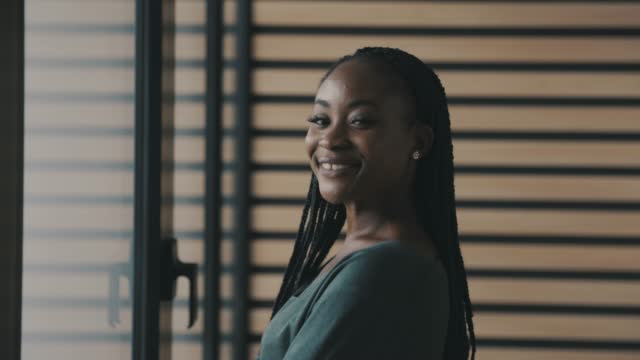 Video portrait of Black businesswoman looking out the window, turning towards camera and smiling joyfully