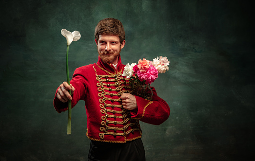 Cavalier and beau. Handsome young man in image of medieval hussar holding flowers isolated on dark blue background. Retro style, comparison of eras concept. Male model wearing hussar uniform.