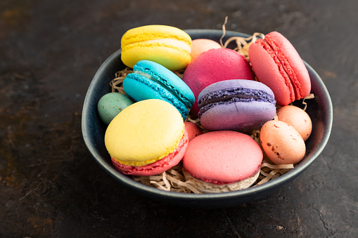 Multicolored macaroons and chocolate eggs in ceramic bowl on black concrete background. side view, close up, still life. Breakfast, morning, concept.