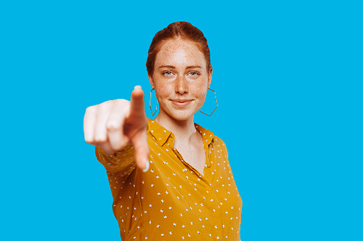 Outgoing carefree young woman points finger directly at camera with positive expression, says I choose you, wears casual outfit, stands over blue wall with blank space area. You are my type.