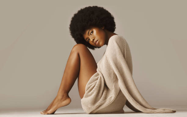 캐주얼 한 겨울 스웨터를 착용하고 맨발로 앉아서 무릎을 꿇고있는 아프로와 함께 아름다운 아프리카 계 미국인 여성. 갈색 배경에 따뜻하고 아늑한 니트웨어를 입고 빛나는 피부를 가진 매력� - african ethnicity beauty curly hair confidence 뉴스 사진 이미지