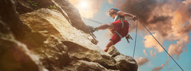 istock Muscular climber man in protective helmet abseiling from cliff rock wall using rope Belay device and climbing harness on evening sunset sky background. Active extreme sports time spending concept. 1402642477