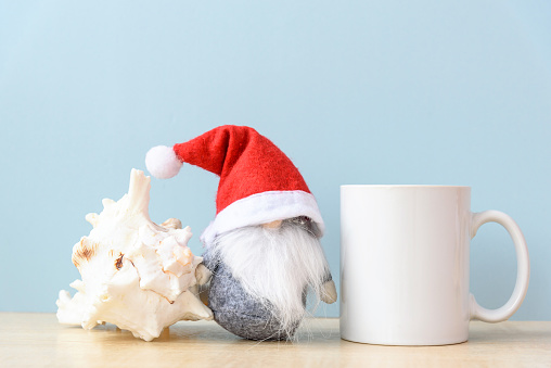 White coffee mug mockup of Christmas in July. Cup, gnome in Santa hat and sea shell