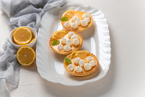 Homemade lemon curd in tartlets with fresh lemon slices, whipped cream and mint on ceramic plate, light concrete background.