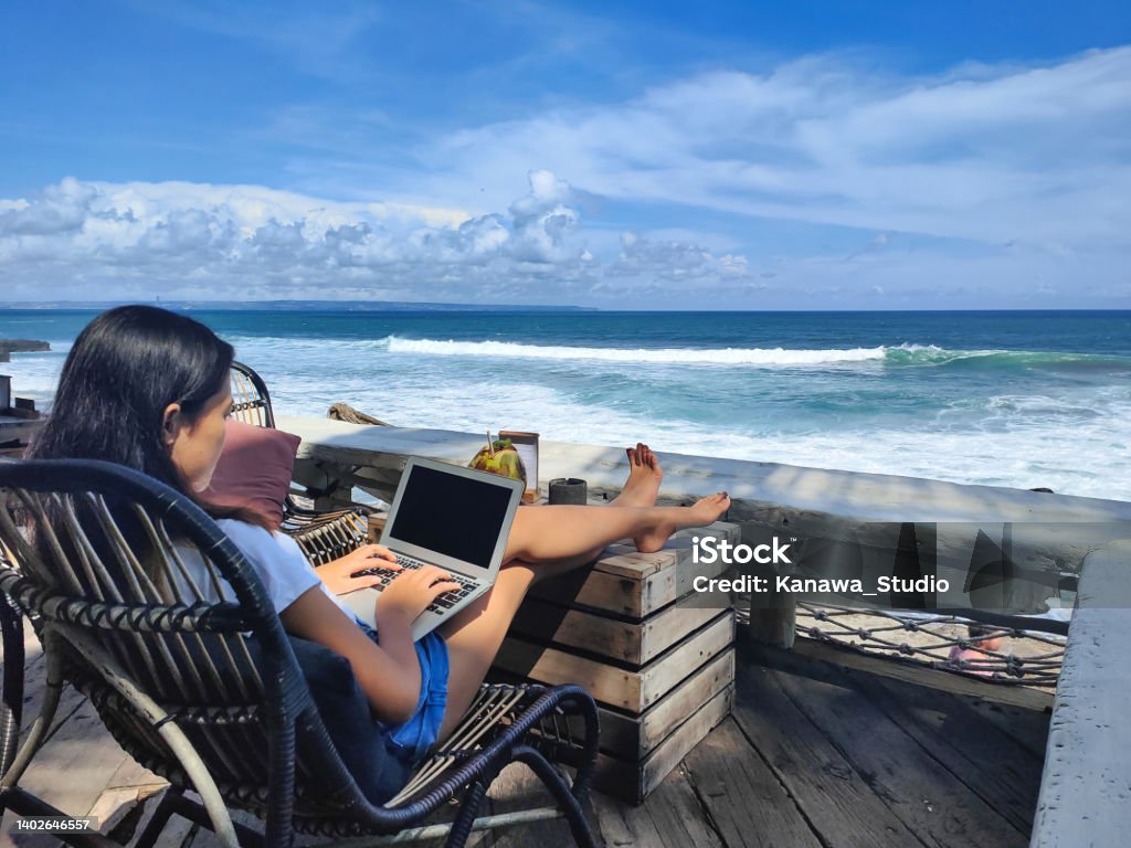 Young Indonesian Woman Working With a Beach View Close-up shot of young female digital nomad working with her laptop, sitting on a chair at a seaside cafe with a beach view. Bali Stock Photo