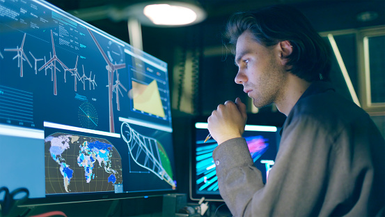 Stock image of a young, long haired man,  working at night in front of a large computer screen showing designs for a wind turbine (wind farm)