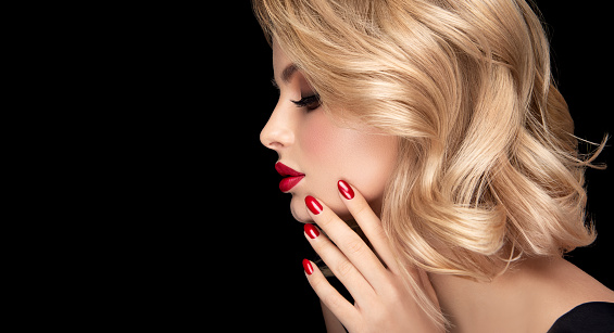 Young woman with perfect wisps on the middle length hairstyle dyed in shades of blonde is demonstrating red manicure on the graceful fingers. Bright makeup with long, blacked eyelashes and vivid red lipstick on the lips.