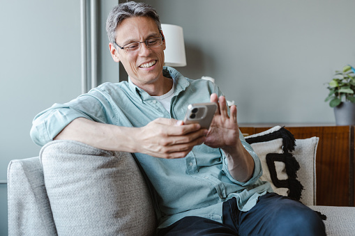 A good-looking man sitting in the living room. He is looking at the mobile phone and consuming internet content.