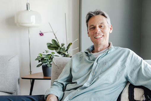 Mature man in casual clothing sitting on the sofa in the living room. He is smiling an looking at the camera.