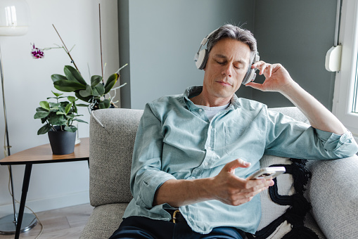 A good-looking man sitting in the living room. He is looking at the mobile phone and consuming internet content. He is listening to music and enjoying the moment