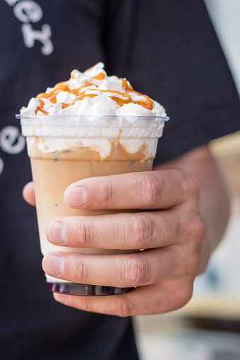 Man holding cold latte with whipped cream. Iced latte decorated with salt caramel topping in hand. Cold drinks. Frozen cappuccino with syrup. Coffee to go. Summer drinks in plastic glass.