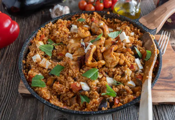 Delicious and healthy dinner or lunch with eggplant and brown rice skillet. Cooked with mediterranean vegetables, feta cheese and spicy chicken breast. Served in a pan on rustic and wooden table background