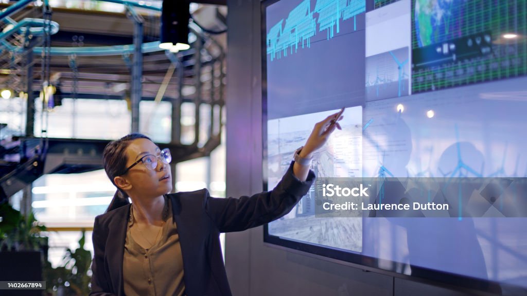 Energy lecture screen Stock photograph of a young Asian woman conducting a seminar / lecture with the aid of a large screen. The screen is displaying data & designs concerning low carbon electricity production with solar panels & wind turbines. These are juxtaposed with an image of conventional fossil fuel oil production. Technology Stock Photo