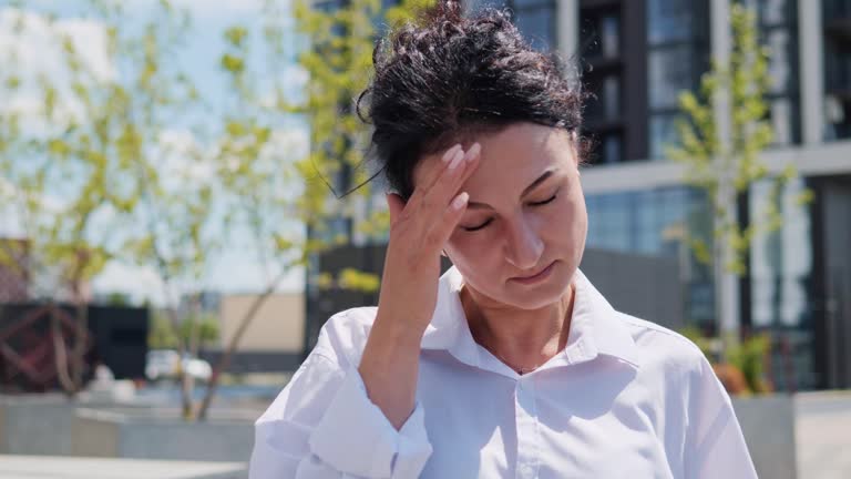 Tired exhausted young adult businesswoman, office worker having headache standing outdoors. Stressed overworked female massages head. Woman feeling terrible pain, migraine headaches, weather changes.