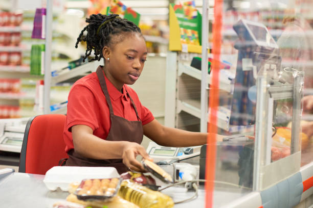 Young black woman in uniform scanning packed food products of buyers Young black woman in uniform scanning packed food products of buyers while sitting by her workplace against displays with shelves cashier stock pictures, royalty-free photos & images