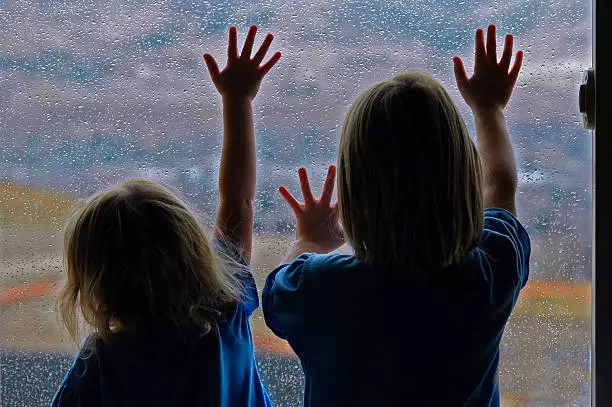 Two little girls standing by window with raindrops on it on a rainy day