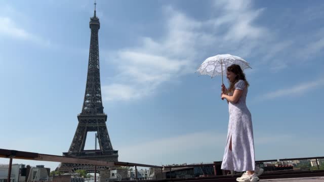 beautiful young teenager girl in paris on the background of the eiffel tower in a long elegant dress in the style of romanticism walks with an umbrella from the sun and smiles at her long blonde hair and around good weather and in the background the eiffe