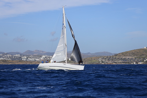 Bodrum,Mugla, Turkey. 10 April 2022: sailor team driving sail boat in motion, sailboat wheeling with water splashes, mountains and seascape on background. Sailboats sail in windy weather in the blue waters of the Aegean Sea, on the shores of the famous holiday destination Bodrum. Men and women on a sailboat in a very cold weather continue to cruise. Sailing crew on sailboat during regatta. Sailing in the wind through the waves at the Sea. Close up of sailing boat, sail boat or yacht at sea with white sails.