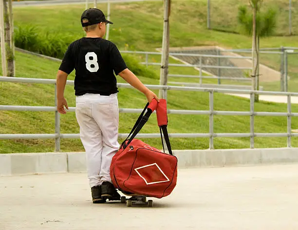 Little boy in baseball uniform using his skateboard to carry his duffel bag
