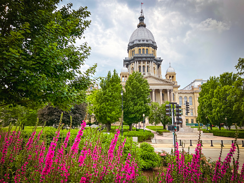 Views of the Illinois State Capitol Building in Springfield, IL, USA as viewed through a blooming formal garden. Dramatic cloudscape and green leafy trees line the perimeter.