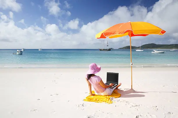 Photo of Woman sitting on a beach towel using laptop under a parasol