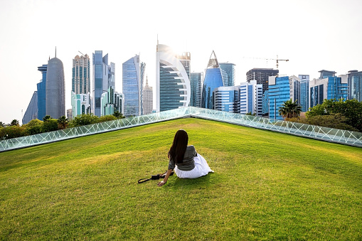 Indian solo traveller in Qatar, Middle East. Young woman sitting on a green lawn in a park with modern skyscrapers of West Bay of Doha Downtown skyline on background