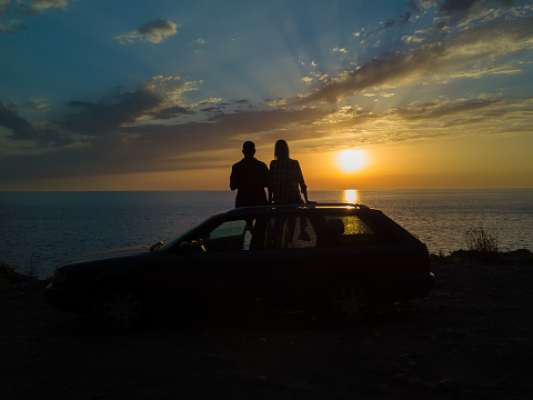 Silhouette of a happy couple sitting on the roof of a car on a cliff in front of the sea with a beautiful orange-blue sky at sunset, view from a drone. Concept of vacations and travel, romance
